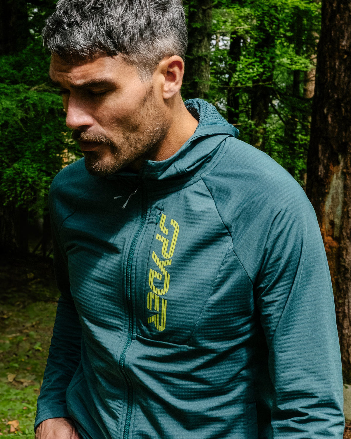 Man wearing a teal jacket with a brand logo in a forest setting