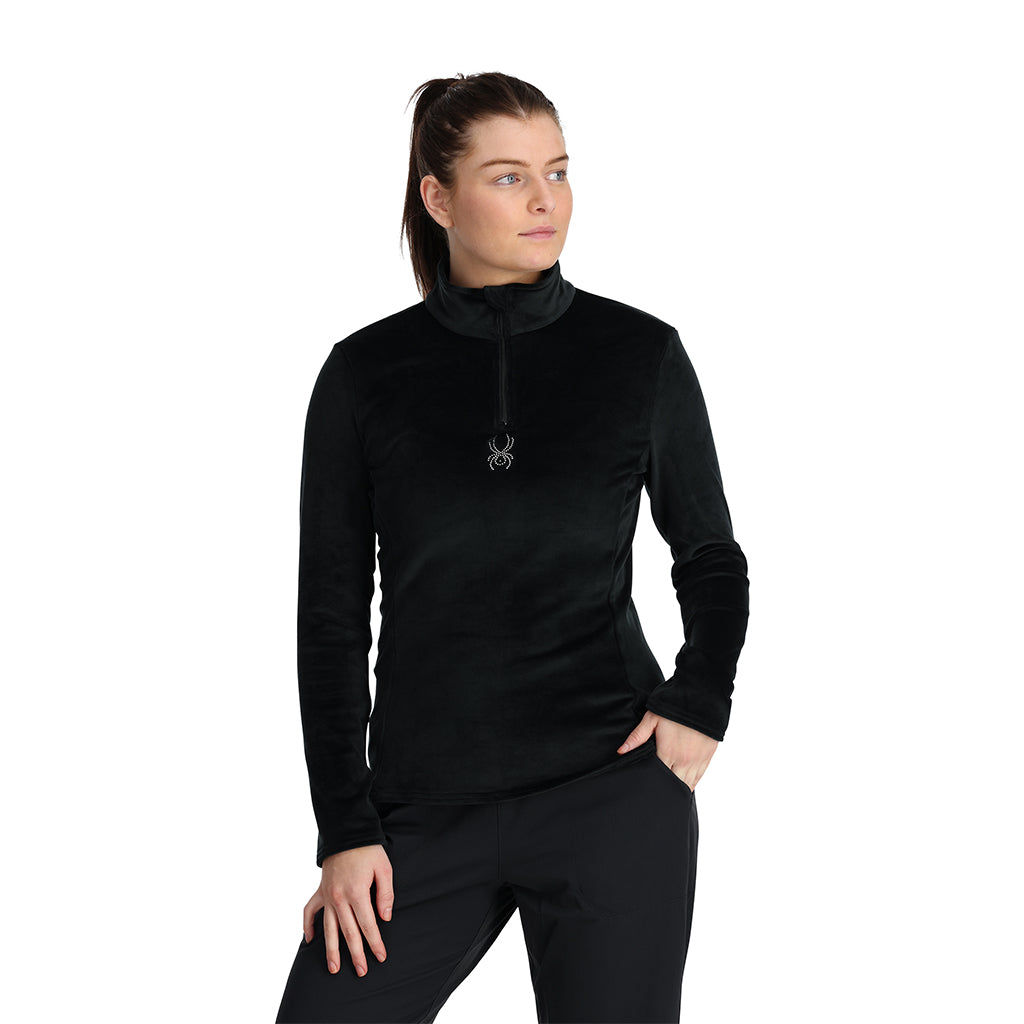 A woman wearing a black Spyder Shimmer Bug half zip pullover with a high neck and a front half zipper.