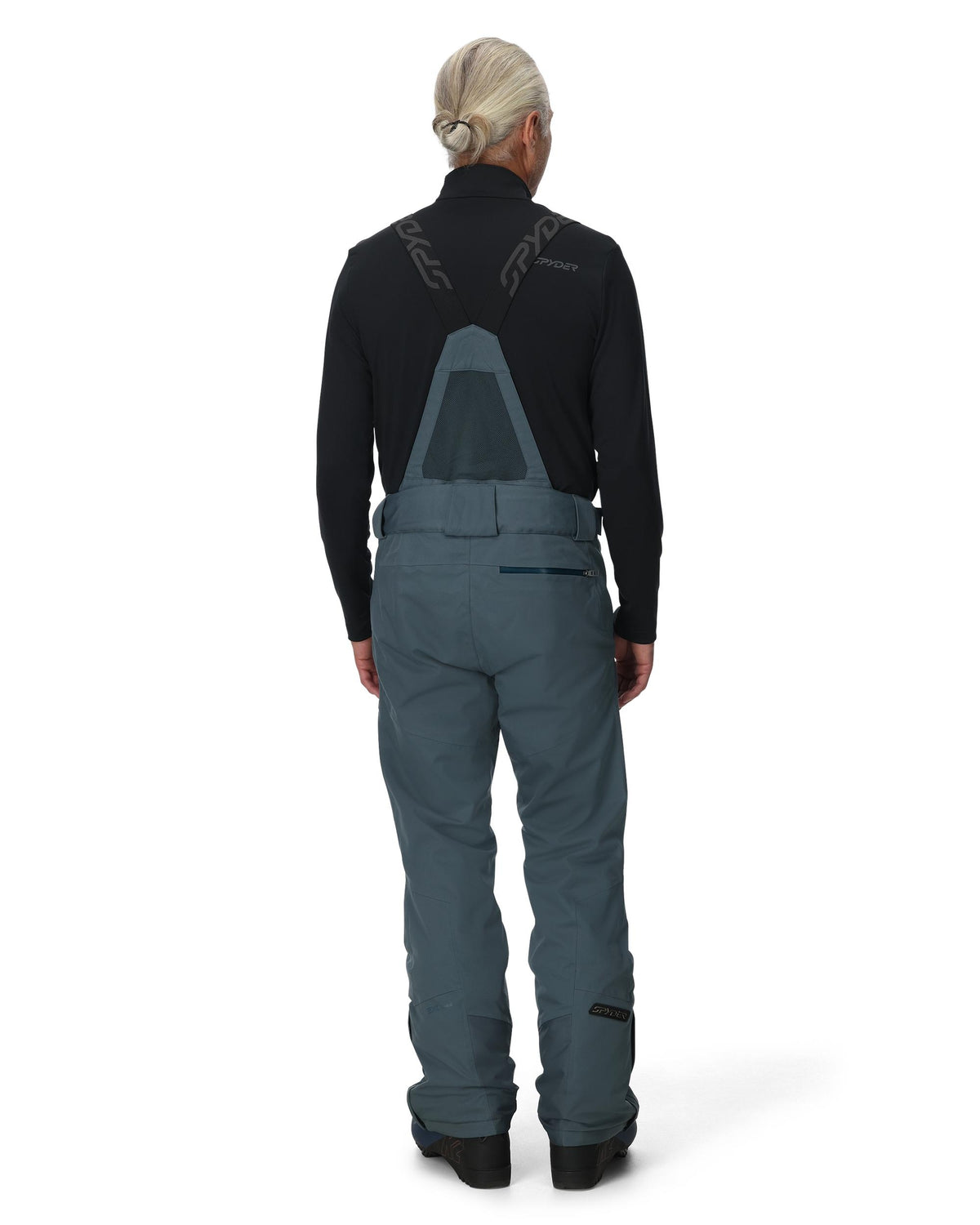 Person wearing a black jacket and teal bib overalls on a white background