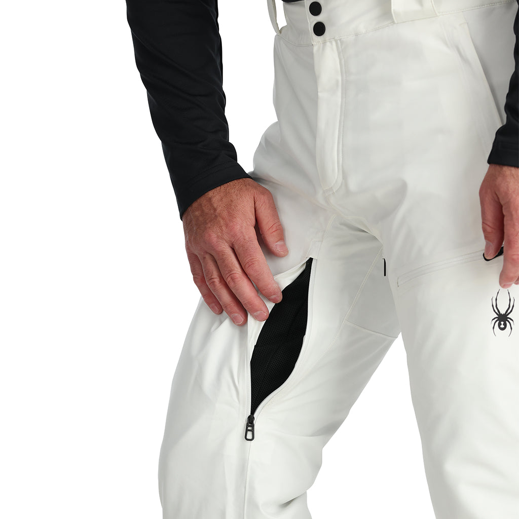 A man standing and wearing white ski pants with adjustable suspenders, a black shirt, and ski boots.
