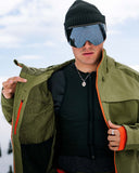 Person wearing a green jacket with orange accents, black beanie, and blue goggles against a snowy background.