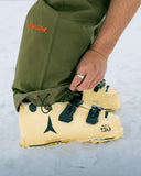 Person wearing a green jacket with a 'Venom' logo, stepping into yellow ski boots on a snowy background.