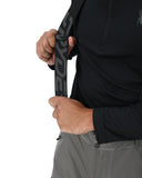 Person holding a black strap with a logo, wearing a black jacket and gray pants on a white background