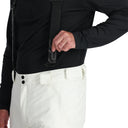 A man standing and wearing white ski pants with adjustable suspenders, a black shirt, and ski boots.