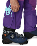 Person wearing purple pants with a blue spider logo, putting on a black and blue ski boot.