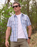 Man wearing a blue plaid shirt over a white t-shirt in a forest setting